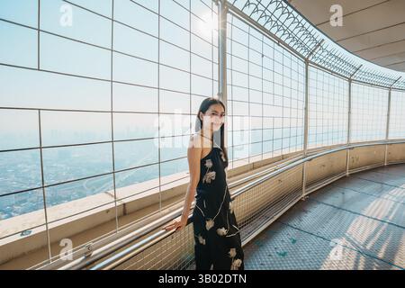 Bella donna asiatica elegante in abito nero che gode di una vista panoramica della città dalla piattaforma di osservazione e di una moderna finestra di vetro nel grattacielo di Bangkok Foto Stock