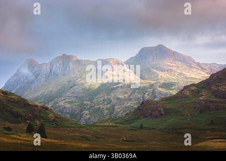 Una mattinata drammatica nel Lake District, dove la luce del sole dorata bagna le aspre pike di Langdale, proiettando una luce serena sul maestoso paesaggio. Foto Stock