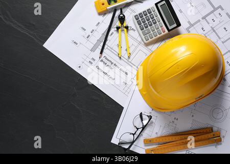 Diversi disegni tecnici, elmetto, matita, divisore, occhiali, righello pieghevole e calcolatrice su tavolo nero, posizione piana. Spazio per il testo Foto Stock