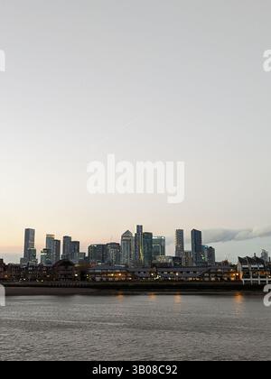Una vista degli alti edifici di Canary Wharf dall'altra parte del fiume tamigi in prima serata contro un cielo grigio chiaro Foto Stock