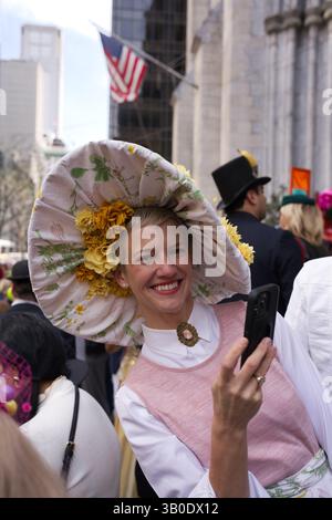 Una donna con un grande cappello floreale scatta una foto con il suo telefono. Indossa un top senza maniche rosa e una camicia con colletto bianco. Lo sfondo è una strada cittadina Foto Stock