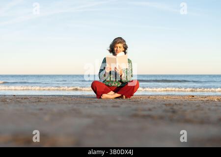 Donna anziana che si gode il suo tempo libero, leggendo un libro accattivante mentre si siede sulla spiaggia al tramonto Foto Stock