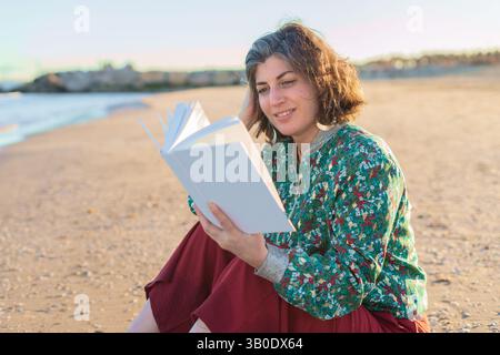Donna matura e felice seduta sulla spiaggia di sabbia, godendo un buon libro mentre si immerge nel caldo tramonto. Indossare abiti casual e sorridere, irradiare Foto Stock