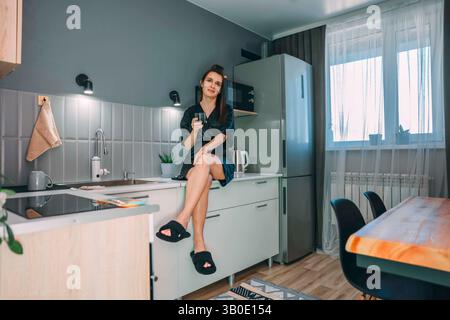 donna seduta sul bancone della cucina in accappatoio nero con cerotti sotto gli occhi sorridenti mentre tiene un bicchiere d'acqua in un moderno e luminoso interno della cucina con morbidi Foto Stock