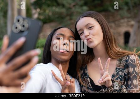 Due giovani donne che creano volti divertenti e segnali di pace mentre scattano un selfie con uno smartphone in un parco vivace, irradiando gioia e amicizia spensierata Foto Stock