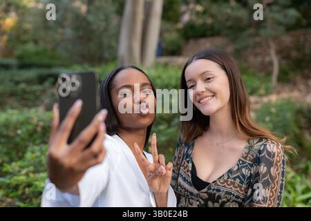 Due amiche stanno facendo volti divertenti e gesti delle mani mentre fanno un selfie con uno smartphone in un parco pubblico Foto Stock