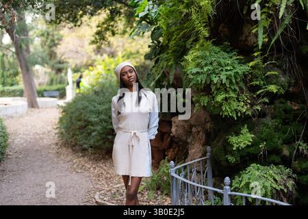 Modella di moda che cammina graziosamente attraverso un lussureggiante giardino botanico, vestito con un elegante abito beige e un accogliente berretto bianco, assaporando il rinfrescante Foto Stock