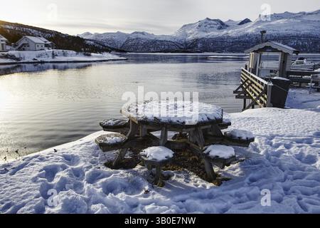 Tavoli e panchine innevati sul lago con montagne sullo sfondo, tranquilla atmosfera invernale, Stravnjunnje, Innhavet, Norvegia, Europa Foto Stock