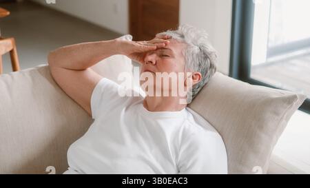 Una donna anziana siede su un divano e tiene la testa tra le mani, ha mal di testa Foto Stock