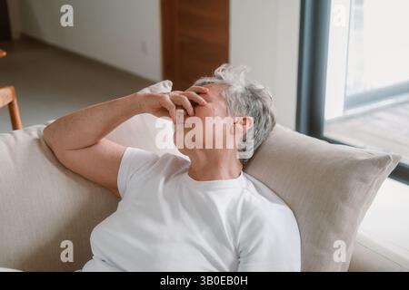 Una donna anziana siede su un divano e tiene la testa tra le mani, ha mal di testa Foto Stock