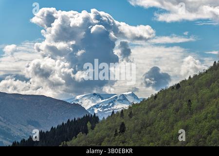 Le cime innevate si innalzano su colline verdeggianti, con soffici nuvole che scivolano attraverso un cielo azzurro, creando un suggestivo panorama naturale. Foto Stock