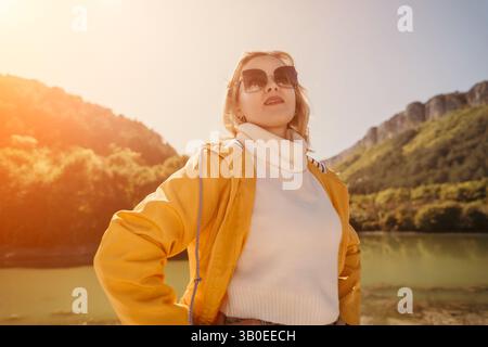 Donna occhiali da sole Mountain Landscape - Una donna che indossa occhiali da sole si erge con sicurezza sullo sfondo di un paesaggio montuoso. Il sole splende Foto Stock