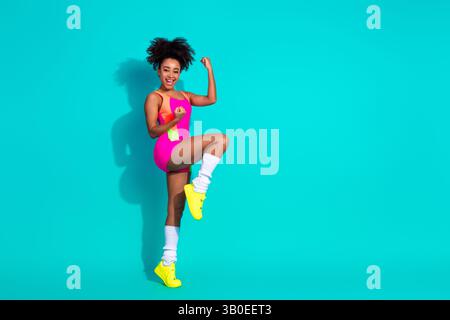 Giovane donna sorridente che indossa un vivace abbigliamento sportivo che mostra forza ed energia su uno sfondo turchese Foto Stock