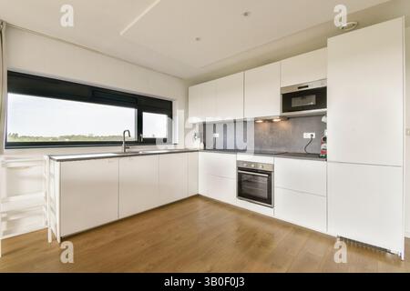 Un'elegante cucina moderna con mobili bianchi, un ripiano grigio e pavimento in legno, con luce naturale che scorre attraverso una grande finestra. Foto Stock