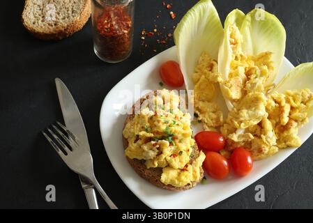 Uova strapazzate con spezie ed erbe aromatiche su croccante pane tostato integrale e verdure in un piatto bianco su sfondo nero. Colazione salutare. Vista dall'alto. Foto Stock
