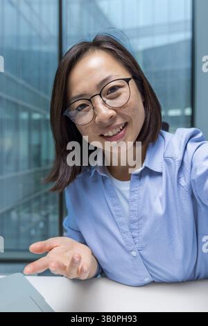 Giovane donna d'affari che utilizza l'app per telefono per videochiamate e la visualizzazione della fotocamera dello smartphone. Lavoratore che guarda la fotocamera sorridendo e parla con colleghi e partner seduti alla scrivania all'interno dell'ufficio. Foto Stock