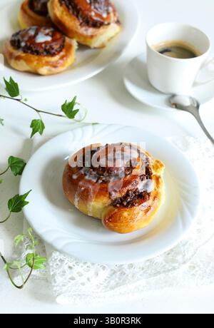 Dolci alla cannella. Rotolo di brioche di noci, decorato con un velo di glassa e una tazza di caffè su sfondo bianco in legno. Lumache di noce Nuss-schnecke. Foto Stock