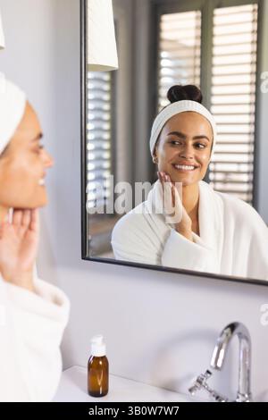 Donna sorridente in accappatoio che tocca la guancia allo specchio del bagno, con biberon in vetro ambrato Foto Stock