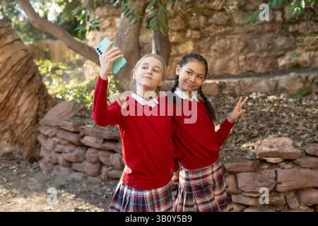 Due studentesse in uniforme scolastica si stanno compiendo un selfie in un parco, abbracciandosi l'un l'altro mentre lampeggiano i segnali di vittoria con le dita e Foto Stock
