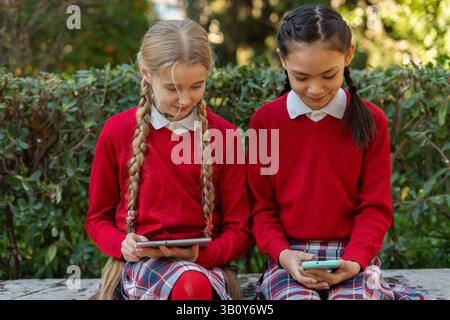 Due ragazze che indossano le uniformi scolastiche stanno usando un tablet e uno smartphone, sedute su una panchina in un parco e si godono la tecnologia durante la pausa Foto Stock