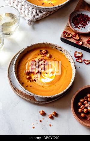Una ciotola di cremosa zuppa di carote guarnita con fette di peperone rosso, ceci croccanti e un turbinio di panna, servita insieme a bicchieri di vino bianco e un PL Foto Stock