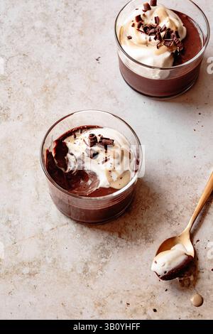 Due tazze di vetro ripiene di mousse al cioccolato ricoperte di panna montata e scaglie di cioccolato, con un cucchiaio a lato. Foto Stock
