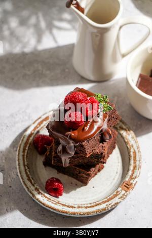 brownie al cioccolato ricoperti di cioccolato fuso lucido e lamponi freschi, serviti su un piatto di ceramica rustica. Il dessert è posto su un li testurizzato Foto Stock
