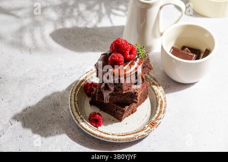 brownie al cioccolato ricoperti di cioccolato fuso lucido e lamponi freschi, serviti su un piatto di ceramica rustica. Il dessert è posto su un li testurizzato Foto Stock