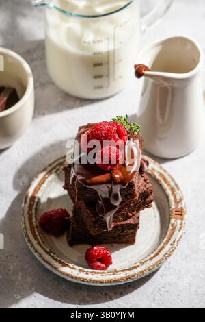 brownie al cioccolato ricoperti di cioccolato fuso lucido e lamponi freschi, serviti su un piatto di ceramica rustica. Il dessert è posto su un li testurizzato Foto Stock