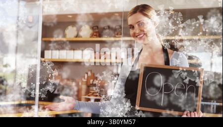 Barista che tiene aperto cartello lavagna dietro il bancone in caffetteria, con decalcomanie virus, vasetti e tazze Foto Stock