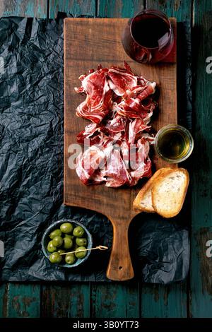 Prosciutto spagnolo affettato finemente Jamon Iberico paleta su tagliere in legno. Bicchiere di vino rosso, olive verdi, olio d'oliva, pane tostato. Vecchio turchese scuro Foto Stock