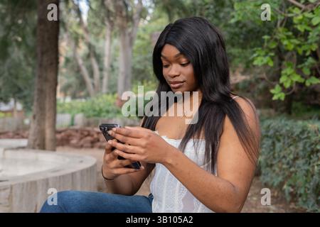 Giovane donna nera seduta tranquilla in un parco, godendo di un momento di relax mentre usa il suo smartphone, circondata dalla bellezza della natura su una soleggiata S Foto Stock