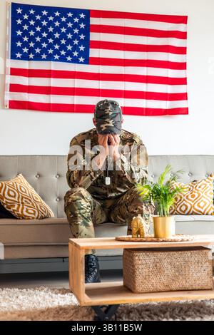 Veterano militare depresso verticale seduto su un divano con bandiera degli Stati Uniti sul muro che copre il viso con le mani che soffrono di stress post-traumatico diso Foto Stock