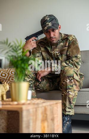 Soldato depresso seduto sul divano di casa, indossando uniforme mimetica e stivali militari, tenendo una pistola vicino alla testa, soffrendo di post-traumatico Foto Stock
