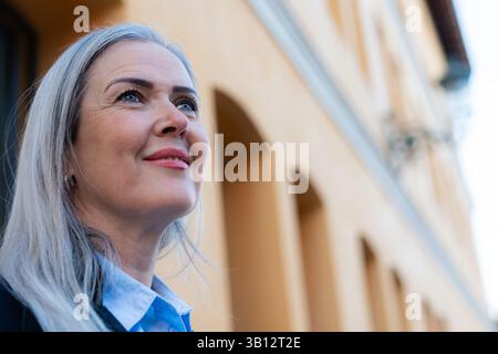 Ritratto di una donna d'affari di successo con i capelli grigi, in piedi con fiducia di fronte a un edificio moderno, sorridente e con un senso di speranza Foto Stock