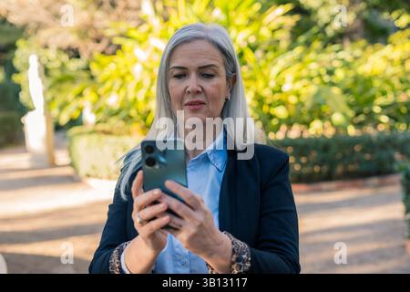 Donna d'affari dai capelli grigi vestita con una giacca, che tiene in mano uno smartphone e si impegna con un'app mentre è circondata da una vegetazione lussureggiante in un parco illuminato dal sole Foto Stock