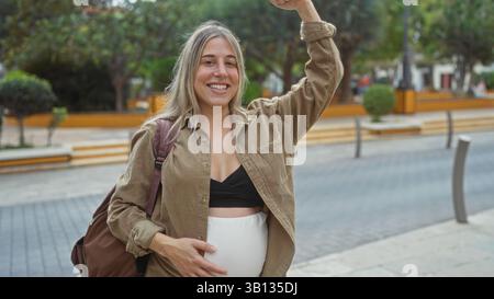Donna incinta che mostra forza in un parco cittadino, sorride con fiducia mentre ammira la sua pancia, incarna l'empowerment e la felicità in una città Foto Stock