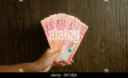 Donna che tiene in mano più banconote del peso filippino su uno sfondo rustico in legno, mostrando banconote in valuta in mano con le dita visibili. Foto Stock