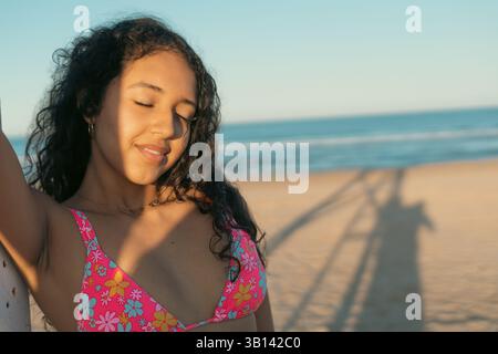 Giovane donna a occhi chiusi che gode di un tranquillo tramonto sulla spiaggia. Indossando un costume da bagno floreale, è rilassata contro un palo con onde e ombre oceaniche Foto Stock