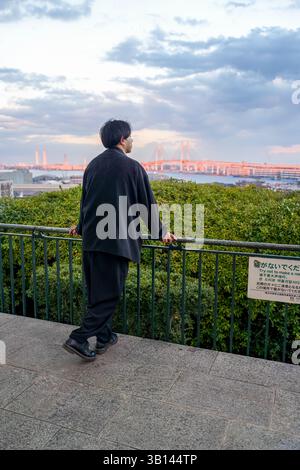 Un uomo giapponese sui vent'anni che indossa una giacca nera si trova a Minato Mirai, Yokohama, prefettura di Kanagawa. Visto da dietro, guarda un ponte lontano Foto Stock