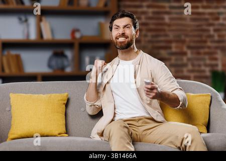 Fan che guardano la partita, celebrano la vittoria, amici entusiasti che guardano la TV, uomo seduto su un comodo divano, pugni sollevati in trionfo mentre guardano un intens Foto Stock