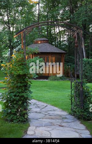 Percorso in pietra ammiraglia grigia e abbronzata attraverso un mandrino in metallo nero ricoperto da Vitis arrampicata - viti e gazebo nel giardino sul retro in estate al crepuscolo. Foto Stock