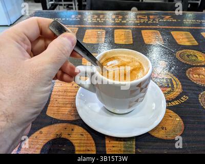 Una persona che mescola un espresso appena estratto in una tazza bianca su un piattino, collocato su un rustico tavolo di legno. Foto Stock