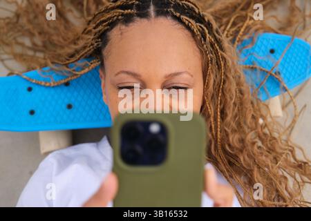Giovane donna con trecce bionde sdraiata sullo skateboard usando il telefono Foto Stock