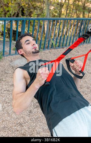 Uomo concentrato che esegue le sospensioni TRX in posizione di parcheggio con le braccia piegate e il torace sollevato, mostrando un controllo del peso corporeo e un coinvolgimento muscolare Foto Stock