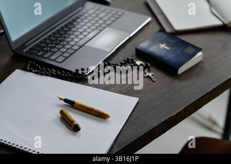 Blocco note aperto con penna vintage, laptop, bibbia e rosario stesi sul tavolo nell'ufficio delle suore, spazio per le copie Foto Stock