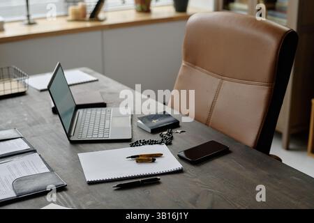 Tavolo in legno scuro con blocco note, penna, bibbia, rosario e dispositivi digitali. Sedia da ufficio in pelle marrone chiaro di fronte al tavolo, spazio fotocopie Foto Stock
