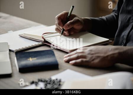 Primo piano medio di un pastore irriconoscibile che tiene in mano la penna durante la scrittura del notebook in ufficio durante la giornata lavorativa Foto Stock