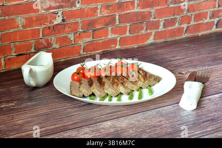 Bistecca di manzo medio rara tagliata a pezzi con pomodori ciliegini e asparagi su un tavolo di legno, accanto a una barca con sugo di carne e posate. Primo piano. Foto Stock