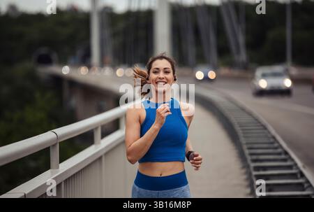 Giovane donna che corre con sicurezza su un ponte, indossa abiti sportivi e cuffie, mentre si allena al mattino mentre le auto passano sullo sfondo Foto Stock